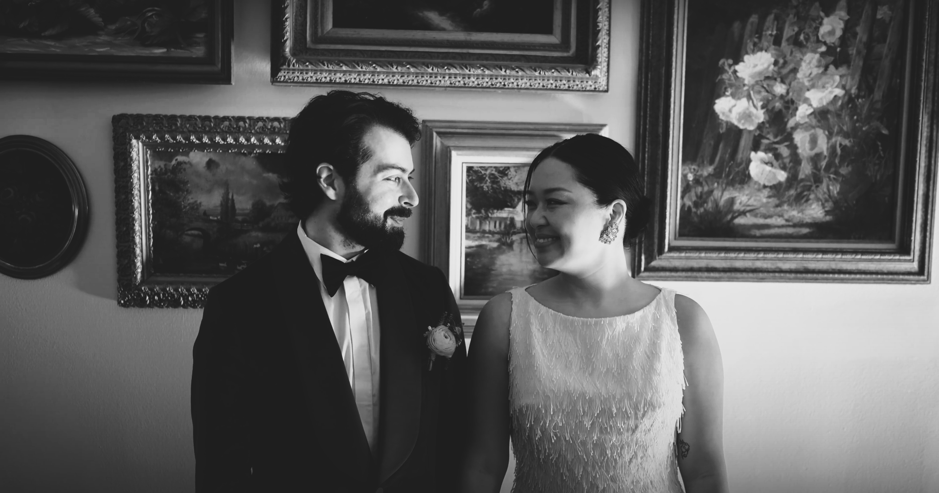 Bride and Groom stand side by side and exchange a loving glance in front of a wall of framed art.